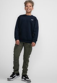 Felpa navy con collo a giro, vestibilità rilassata e dettaglio del logo. Pantaloni cargo verde oliva con tasche, abbinati a sneakers nere e bianche.