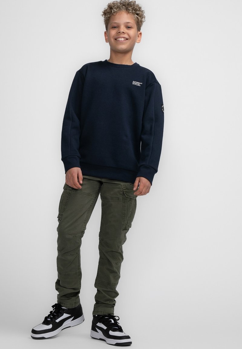 Felpa navy con collo a giro, vestibilità rilassata e dettaglio del logo. Pantaloni cargo verde oliva con tasche, abbinati a sneakers nere e bianche.
