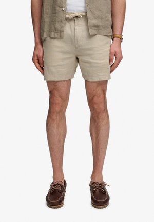 Man draagt beige linnen short met trekkoord, bruine leren bootschoenen, een bruin kort mouw overhemd en een horloge aan de linkerpols.