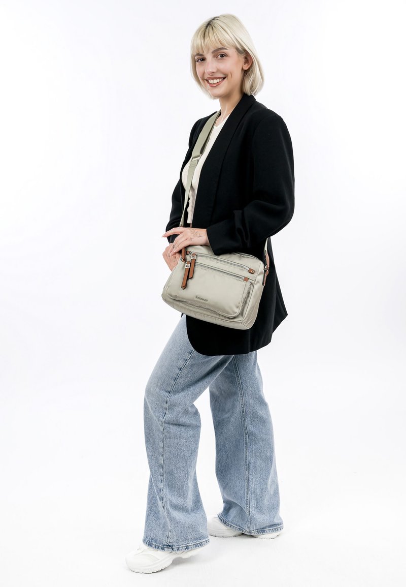 Světlě šedá crossbody taška s texturovaným povrchem, s hnědými koženými detaily a zipovým kováním. Model má na sobě džíny s rozšířenými nohavicemi a černý blazer.