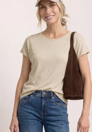 Lächelnde Frau trägt beige kurzärmliges Oberteil, blaue Jeans, beige Kappe und eine braune Veloursleder-Schultertasche.