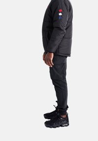 Mann, der seitlich steht und eine schwarze Steppjacke mit rot-weißen-blauen Blumen an den Ärmeln, schwarze Cargo-Hosen und schwarze Sportschuhe trägt.