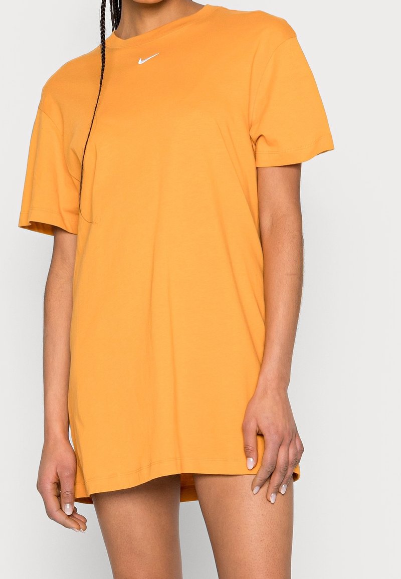 Person klädd i en överdimensionerad orange Nike-t-shirt som klänning, stående med en hand vilande på sin lår mot en enfärgad bakgrund.