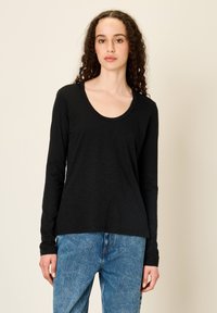 Koolikate juustega naine, kes kannab musta pika varrukaga särki ja siniseid teksaseid, ühtlase beežitaustaga.