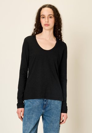 Donna con capelli ricci che indossa una maglietta nera a maniche lunghe e jeans blu, davanti a uno sfondo beige uniforme.