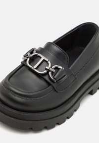 Mocasín de cuero negro con una suela gruesa y texturizada. Presenta un detalle de cadena plateada a lo largo del empeine y costuras decorativas en la parte superior.