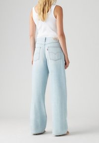 Mujer vistiendo unos pantalones vaqueros de pierna ancha azul claros y una camiseta sin mangas blanca, de pie contra un fondo gris liso, de espaldas.