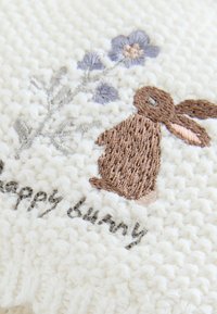 Geborduurde stof met een bruine konijn en bloemmotief, tegen een gestructureerde witte achtergrond. Tekst luidt "happy bunny".