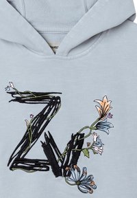 Hellblaue Kapuze mit gesticktem "Z"-Design, umrandet in Schwarz und mit bunten floralen Akzenten. Die Jacke zeigt eine weiche Textur und einen modernen Schnitt.