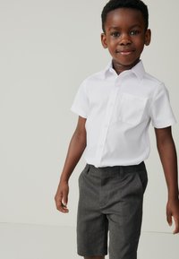 Weißes kurzärmeliges Hemd zum Knöpfen mit Brusttasche, kombiniert mit anthrazitgrauen Shorts. Glatter Stoff, maßgeschneiderte Passform und dezente Details.