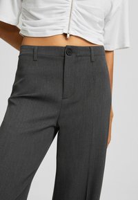 Žena v tmavě šedých kalhotách na míru a bílém zkráceném topu s předním zipem, zobrazená od pasu po střed stehna.