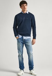 Hombre vistiendo un sudadera azul marino sobre una camisa azul clara, jeans desgastados y zapatillas blancas, de pie con una mano en el bolsillo contra un fondo gris.