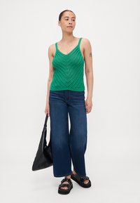 Top sin mangas de punto verde con escote en V y patrón de chevron, combinado con jeans de pierna ancha azules y sandalias de plataforma negras, sosteniendo un bolso negro.