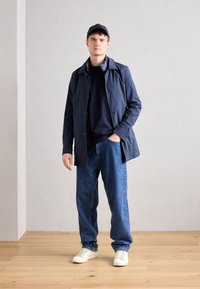 Giovane uomo in cappello blu navy, giacca blu navy, camicia scura, jeans larghi blu e sneakers bianche, in piedi su un pavimento di legno contro un muro semplice.