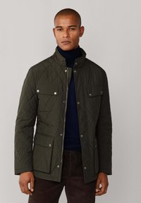 Oliven zelena jakna s kvadratnim uzorkom, s pritiskujućim gumbima, patentnim zatvaračem, dva prednja džepa i stojećim ovratnikom, uparena s mornarskom dolčevitom.