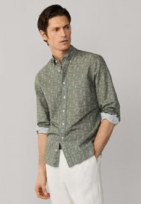 Muž s vyhrnutými rukávy v zelené vzorované košili na knoflíky a bílých kalhotách, stojící s jednou rukou v kapse, hledící doleva.
