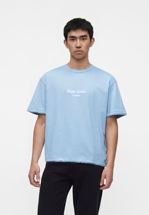 Yngre mand iført lyseblå Pepe Jeans London t-shirt og sorte bukser, stående mod en ensfarvet hvid baggrund.