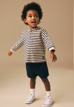 Mladé dieťa s kučeravými vlasmi, oblečené v pruhovanom tričku s dlhými rukávmi, námorníckych šortkách, bielych ponožkách a bielych teniskách, stojace s rozpaženými rukami.