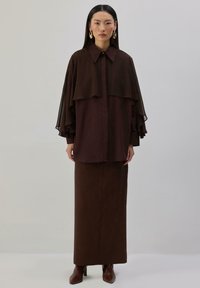 Woman standing wearing a dark brown long skirt, matching layered loose blouse with wide sleeves, gold hoop earrings, and brown heeled boots.