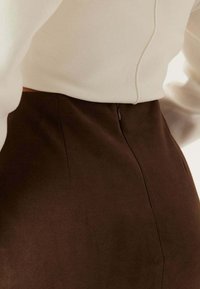 Jupe en daim marron au design ajusté, comprenant une fermeture éclair au dos et des détails de couture. Le haut est de couleur crème avec une finition texturée.