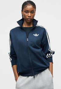 Frau mit geflochtenem Haar, die eine marineblaue Adidas-Jacke mit Reißverschluss und weißen Streifen an den Ärmeln sowie graue Hosen trägt, steht mit den Händen in den Taschen.