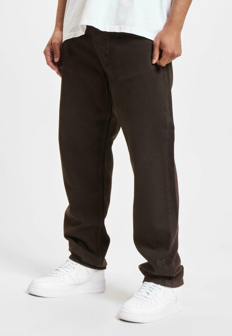 Personne portant un pantalon ample marron foncé, des baskets blanches et une chemise blanche, debout devant un fond uni.