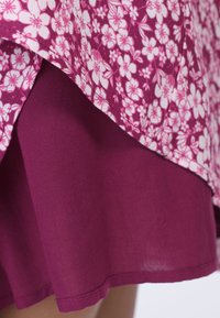 Lila Rock mit einem blumigen Überzug in verschiedenen Rosa-Tönen. Der Stoff scheint leicht und hat eine glatte Textur, mit einem schichtigen Design.