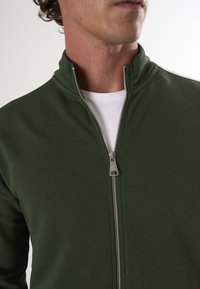 Dunkelgrüne Zip-Jacke mit hohem Kragen und silbernem Reißverschluss, mit einer weißen Innenschicht, die am Ausschnitt sichtbar ist. Glatte Stofftextur.