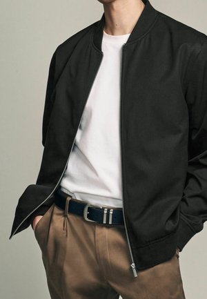 Personne portant une veste zippée noire par-dessus un t-shirt blanc avec un pantalon beige et une ceinture noire, les mains dans les poches, debout devant un fond uni.