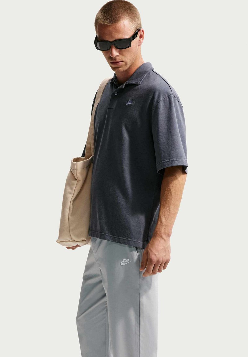 Uomo che indossa una polo Nike grigio scuro, pantaloni Nike grigio chiaro, occhiali da sole neri e che porta una borsa tote beige sulla spalla.