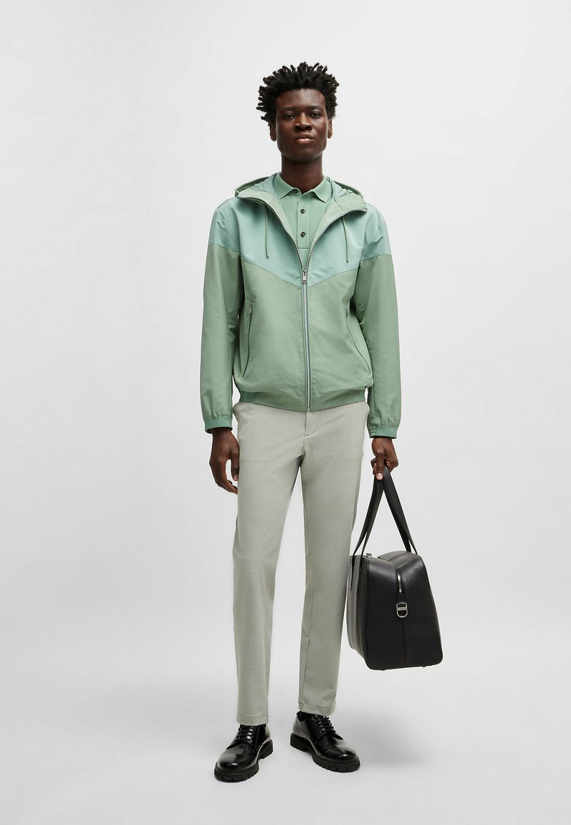 Hellgrüne Zip-Jacke mit Kapuze, die ein Chevron-Design aufweist, kombiniert mit einem hellgrünen Button-Up-Hemd und hellen Hosen. Schwarze Tasche.