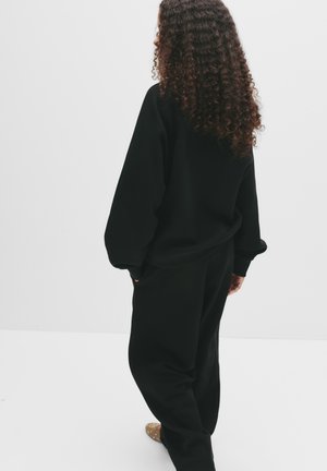 Felpa nera con spalle scese e vestibilità ampia, abbinata a pantaloni neri coordinati. Capelli ricci visibili da dietro.