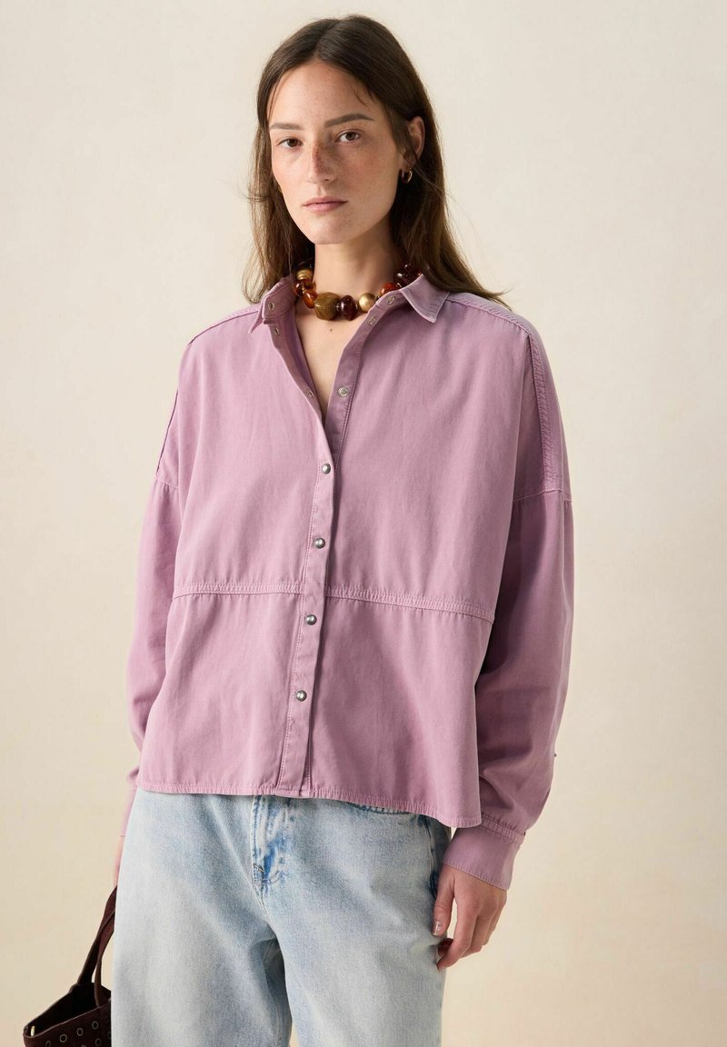 Long-sleeve, button-up shirt in light pink fabric with a relaxed fit, featuring a yoke seam and silver button accents; paired with light blue jeans.