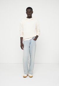 Lys beige struktureret sweater, hvidt undershirt, lyseblå løsthængende jeans og creme sneakers med brune såler. Model poserer foran en ensfarvet baggrund.