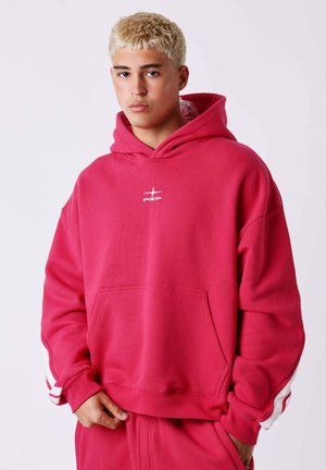 Jeune homme aux cheveux blonds courts portant un sweat à capuche rouge vif avec des bandes blanches sur les manches et un pantalon rouge assorti, debout devant un fond uni.