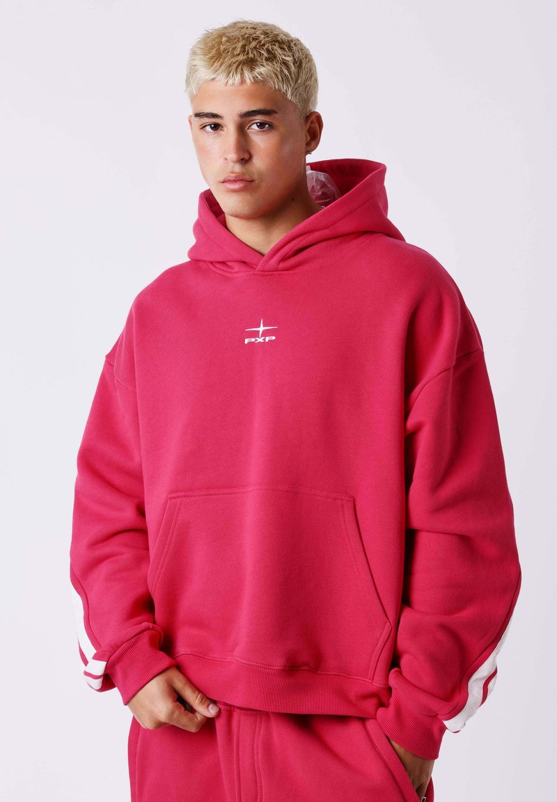 Jeune homme aux cheveux blonds courts portant un sweat à capuche rouge vif avec des bandes blanches sur les manches et un pantalon rouge assorti, debout devant un fond uni.