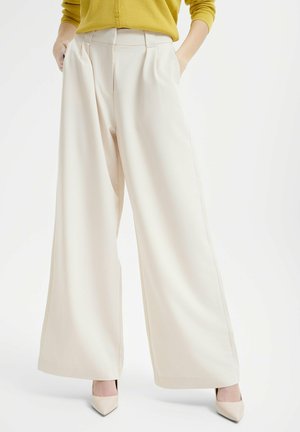 Helle, weit geschnittene Hose in Creme mit Falten und seitlichen Taschen. Der Stoff hat eine glatte Textur und wird kombiniert mit beigen, spitzen Absatzschuhen.