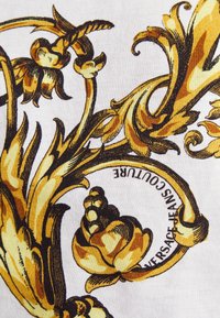 Květinový vzor s použitím zlatých a hnědých odstínů na bílém pozadí, s detailními ilustracemi listů a květin, a textem "VERSACE JEANS COUTURE."