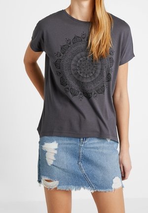 Camiseta de algodón gris con un estampado de mandala negro, combinada con una falda de denim desgastada con dobladillo deshilachado y detalles rasgados.