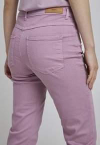 Liila denimist püksid sileda tekstuuriga, millel on tagakotid ja nahklabel vööl, kohandatud kehaõpetusele.