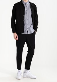 Giacca in denim nera, camicia a bottoni leggera con motivo, pantaloni neri a sigaretta e sneakers bianche con calze. Texture lisce e vestibilità su misura.