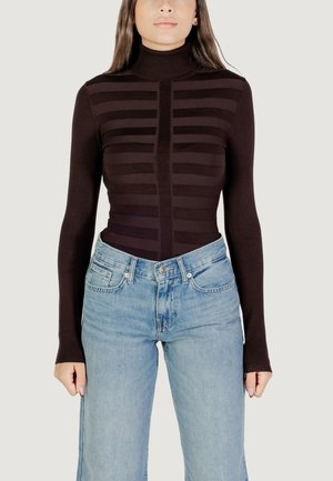 Mujer con un suéter de cuello alto de rayas marrón oscuro y jeans de cintura alta azul claros, de pie contra un fondo sencillo.