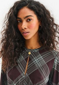 Brązowo-zielona sukienka w kratę z dekoltem typu keyhole i rozkloszowanymi rękawami. Charakterystyczny złoty naszyjnik z zawieszką. Teksturowany, lejący się materiał.