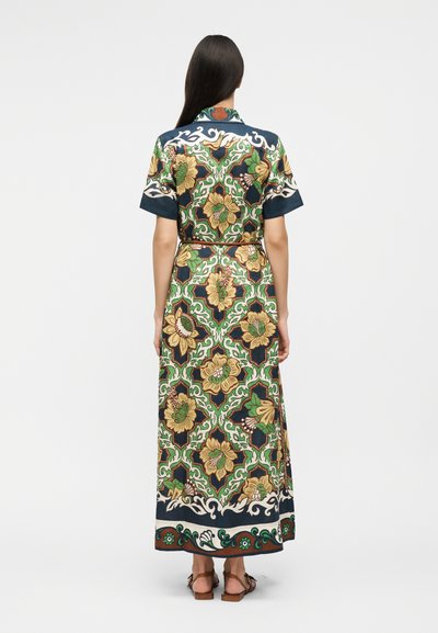Γυναίκα με μακριά σκούρα μαλλιά που φοράει μακρύ φόρεμα με κοντά μανίκια και ζώνη, με πράσινο, χρυσό και ναυτικό μπλε λουλουδάτο σχέδιο, στέκεται με την πλάτη προς τον θεατή.