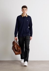 Polo a maniche lunghe blu scuro con colletto beige, abbinato a jeans scuri. Una borsa a tracolla in pelle marrone e sneaker bianche completano il look.