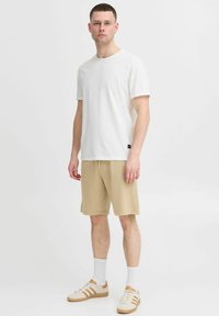 T-shirt blanche à manches courtes associée à un short beige. Des chaussettes blanches et des sneakers beiges avec des accents marron complètent la tenue. Design simple et décontracté.