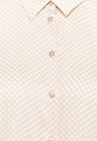 Cream button-up shirt with a light polka dot pattern, featuring a classic collar and five buttons along the front. Smooth fabric texture.