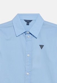 Ανοιχτό μπλε πουκάμισο με ρίγες, κουμπωτό με μύτη γιακά, δύο κουμπιά και σκούρο μπλε ταμπελάκι με το λογότυπο Guess στο αριστερό μέρος του στήθους. Λείο υλικό.