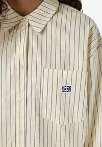 Kollane triibuline nööpidega särk valgete nööpidega, rinna tasku, millel on logo, ja klassikaline krae. Pehme kangas, millel on vertikaalsed jooned.