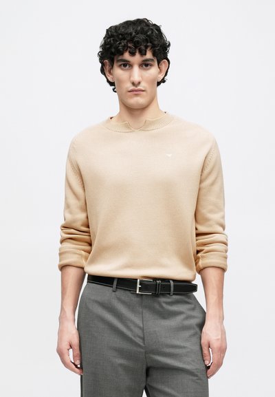 Jeune homme aux cheveux bouclés et foncés, portant un pull en maille beige et un pantalon gris avec une ceinture noire, debout devant un fond uni.